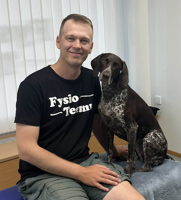 Fysioterapeutti Teemu Uusitalo ja Papu-koira Teemun vastaanotolla Sastamalassa. Teemu ja Papu istuvat fysioterapeutin hoitopöydällä ja poseeraavat vierekkäin kameralle. Teemulla on päällään FysioTeemun logolla varustettu t-paita ja iloinen hymy huulilla. Papu katsoo kameraan rauhallisesti pää kallellaan.